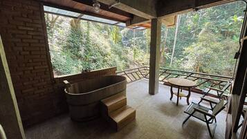 Un baño con una bañera de madera, una mesita pequeña y sillas, y vistas a un jardín exuberante.