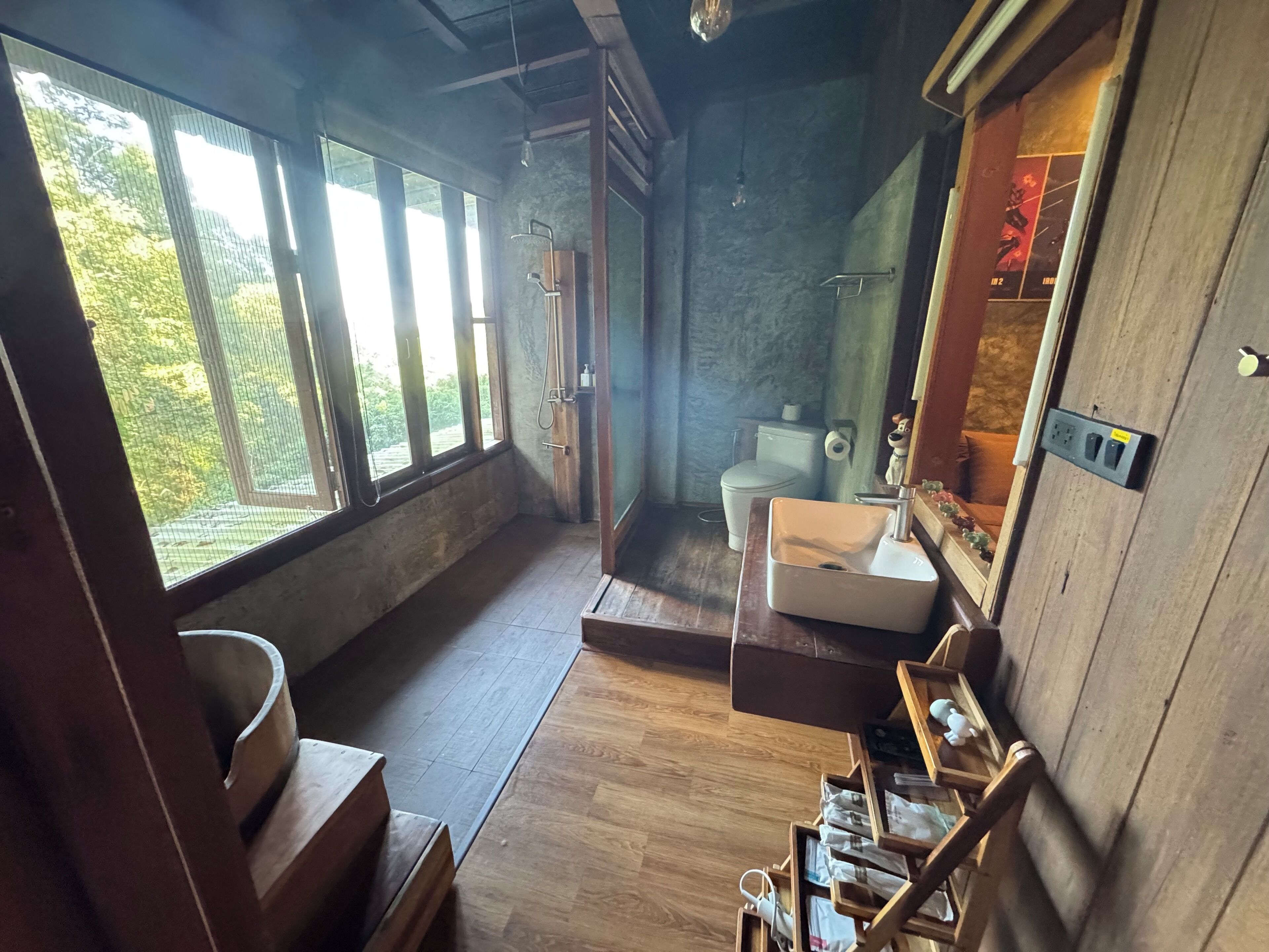A rustic bathroom with a wooden sink, a toilet, and a shower. Large windows provide natural light and a view of greenery outside.