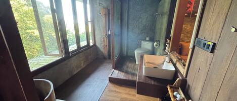 A rustic bathroom with a wooden sink, a toilet, and a shower. Large windows provide natural light and a view of greenery outside.