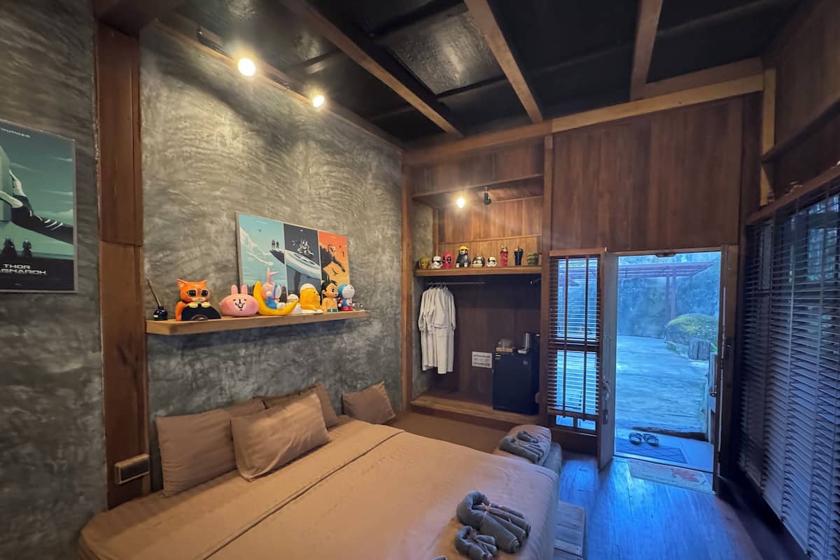 A bedroom with a bed, pillows, a shelf with plush toys, a painting, and a door leading to a balcony.