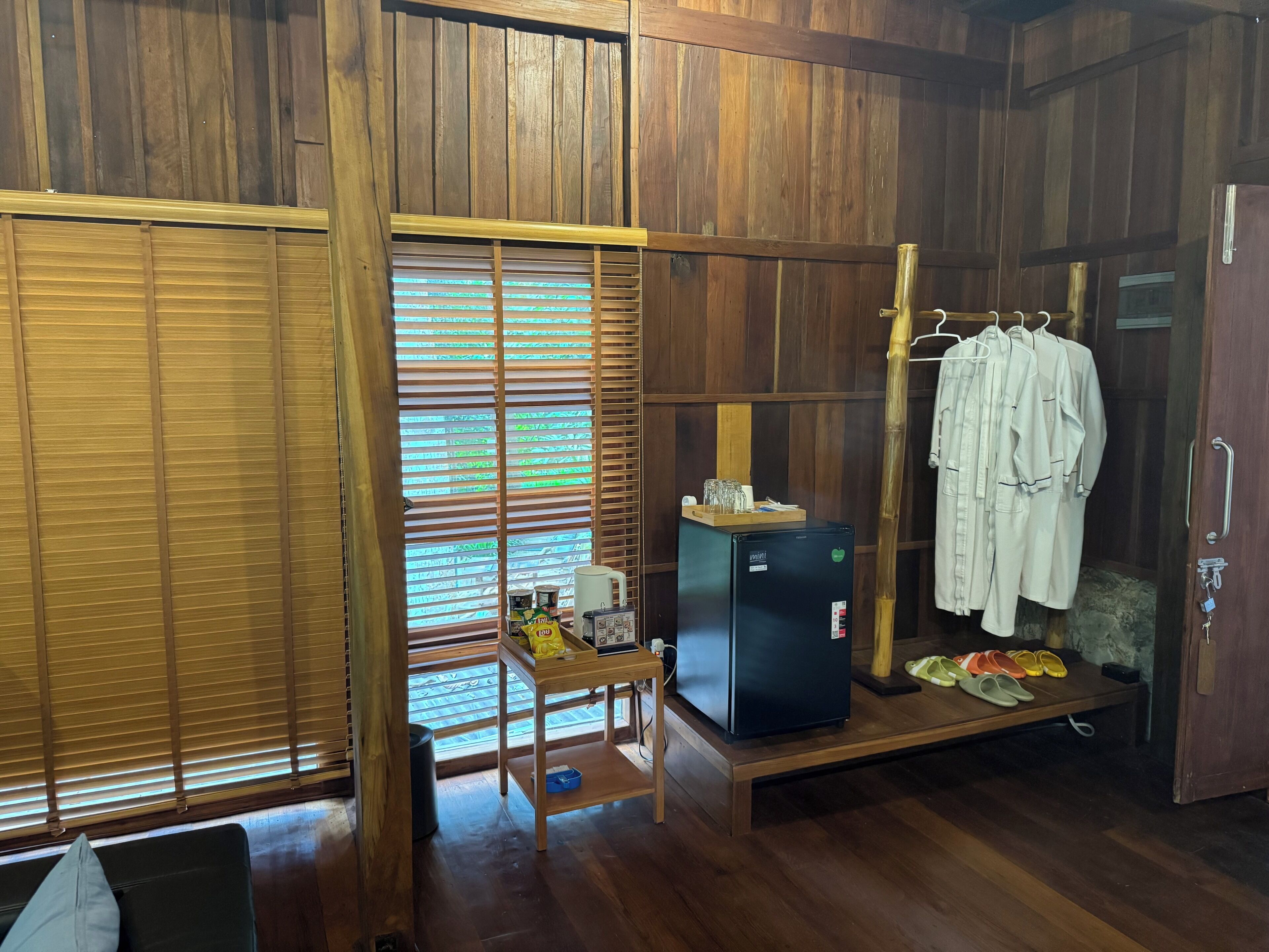 A room with wooden walls, a small table with a refrigerator, a bench, and a clothing rack.