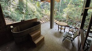 A room with a large bathtub, a small table, and a chair. There is a view of a forest through large windows. - Mii Paa Aii  (Mae On)