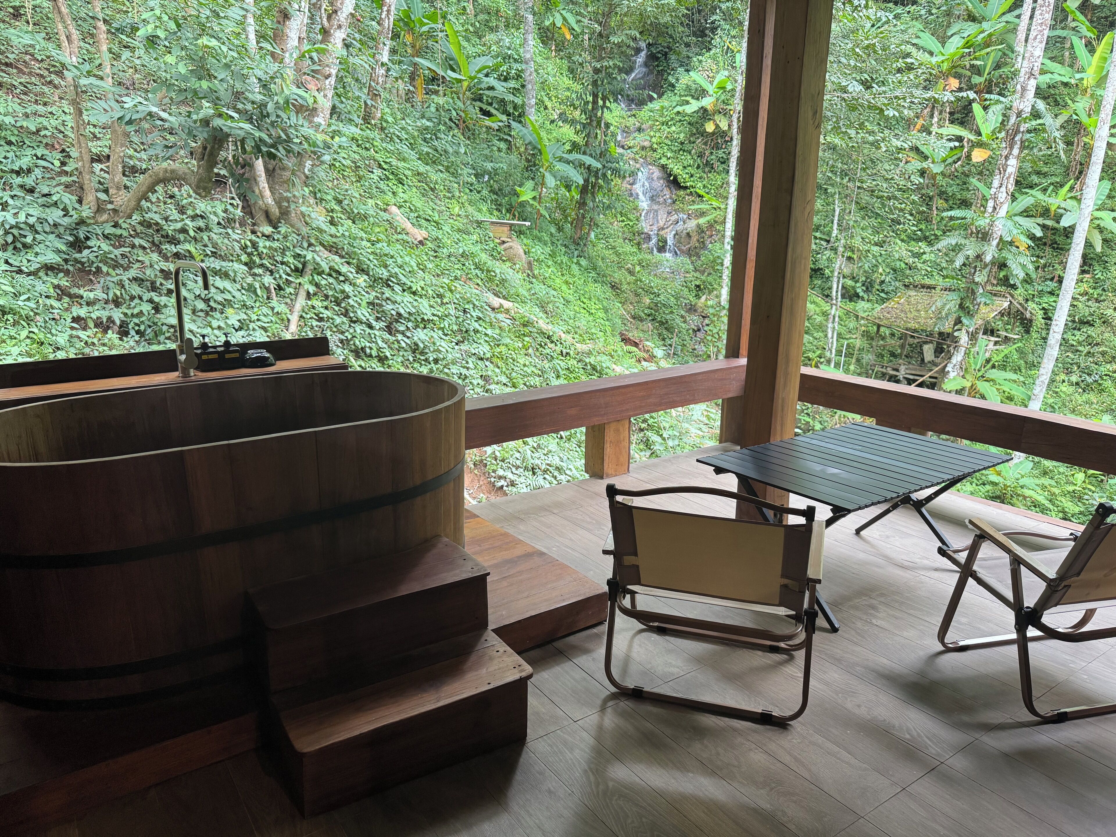Ein Holz-Whirlpool mit Stufen, einem Tisch und zwei Stühlen auf einer Holzterrasse mit Blick auf einen üppigen Wald und einen Wasserfall.