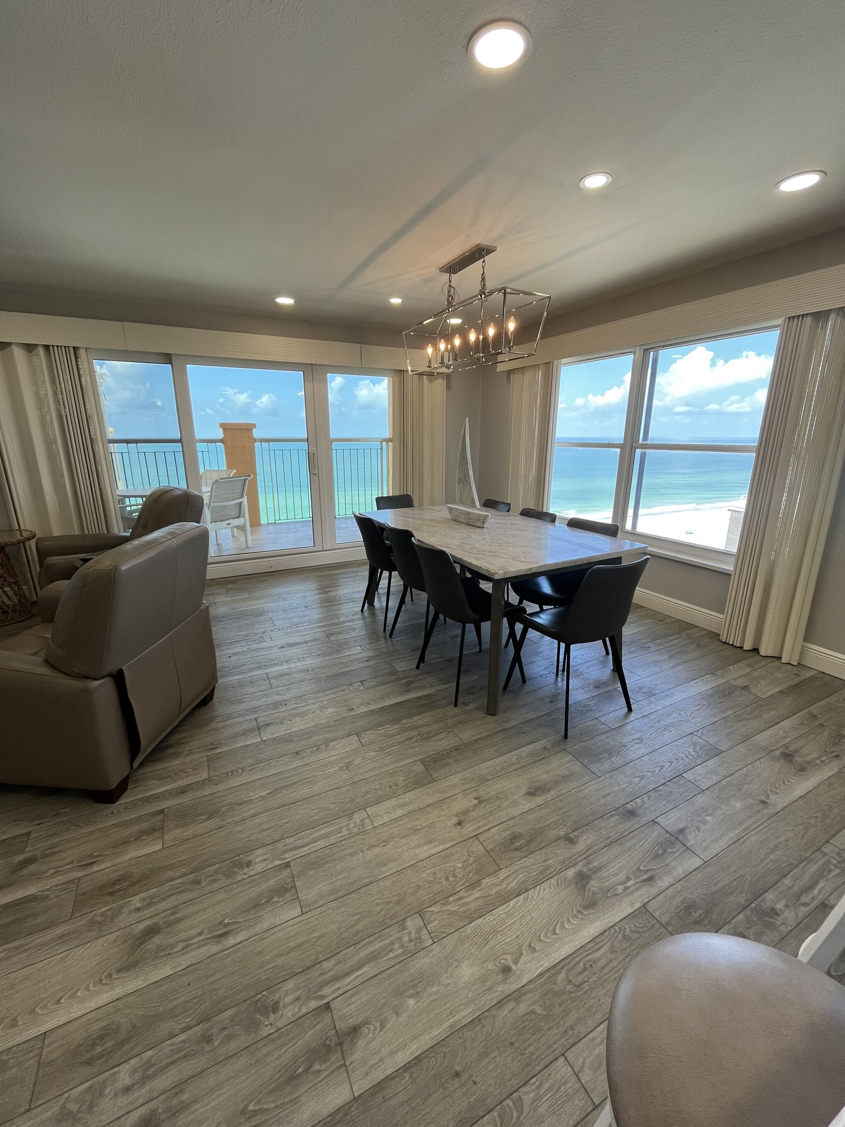 A spacious room with a large dining table, multiple chairs, a sofa, and a view of the ocean through large windows.