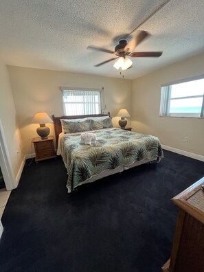 A bedroom with a bed, two bedside tables with lamps, a ceiling fan, a window with blinds, and a view of the outdoors.
