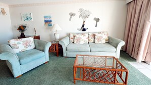 A living room with two light blue sofas, a glass coffee table, and a wall with decorative elements including a palm tree and a guitar.
