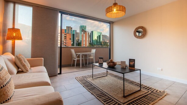 Una sala moderna con un sofá, una mesa de comedor y vistas a un paisaje urbano a través de amplios ventanales.