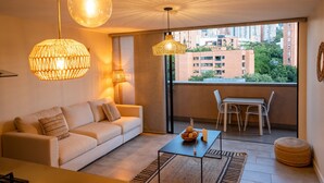 Una sala de estar moderna con un sofá, una mesa de comedor y sillas. Hay luces colgantes y una vista de edificios a través de un ventanal grande.