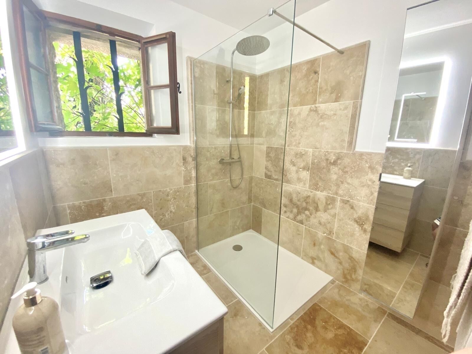 Une salle de bain dotée d’une douche à parois de verre, d’un lavabo blanc avec un miroir et d’une fenêtre donnant sur un paysage verdoyant.