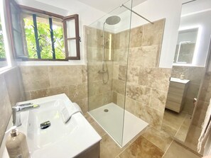 Ein Badezimmer mit einer gläsernen Duschkabine, einem weißen Waschbecken mit Spiegel und einem Fenster mit Blick auf Grünflächen.