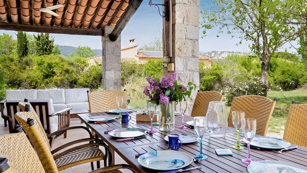 An outdoor dining area with wicker chairs and a wooden table set for a meal, overlooking a scenic landscape.
