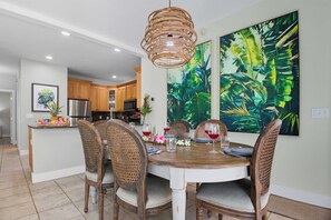 Uma área de refeições com uma mesa redonda de madeira, cadeiras de vime e um quadro grande com tema tropical na parede.