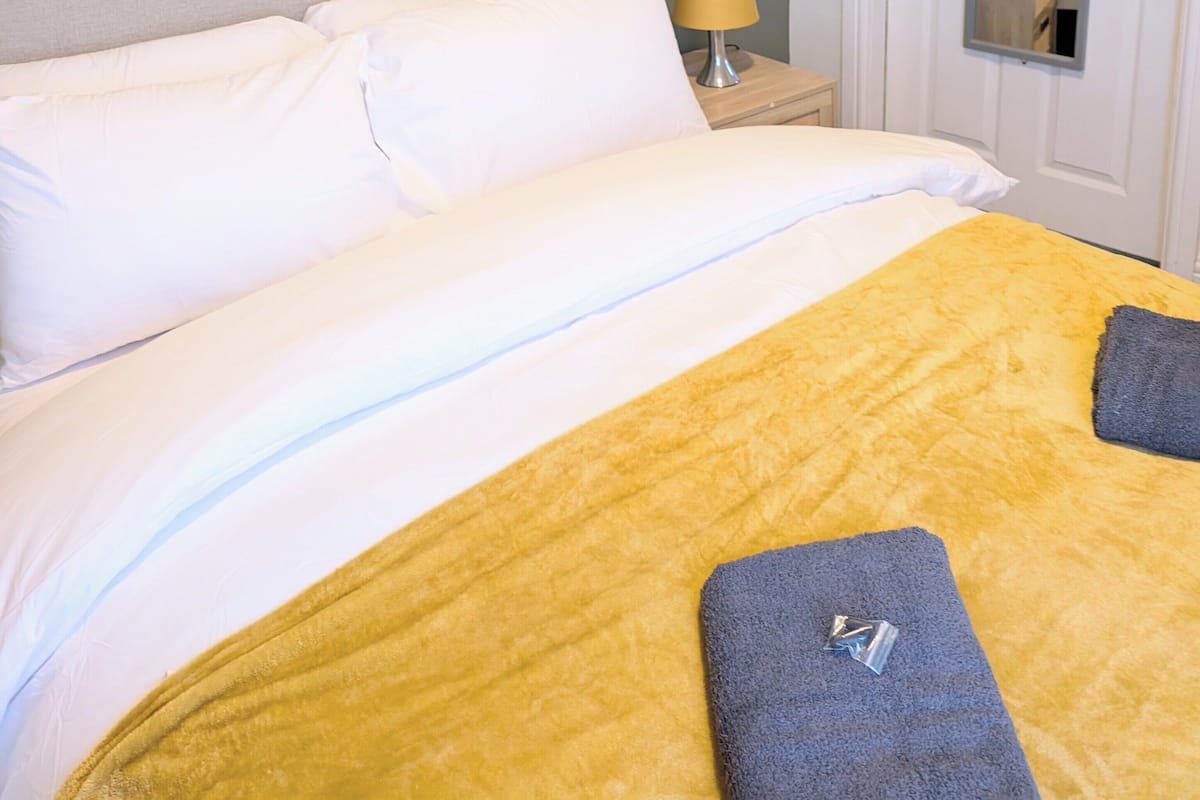 A neatly made bed with white and yellow bedding, a bedside lamp, and a framed artwork on the wall.