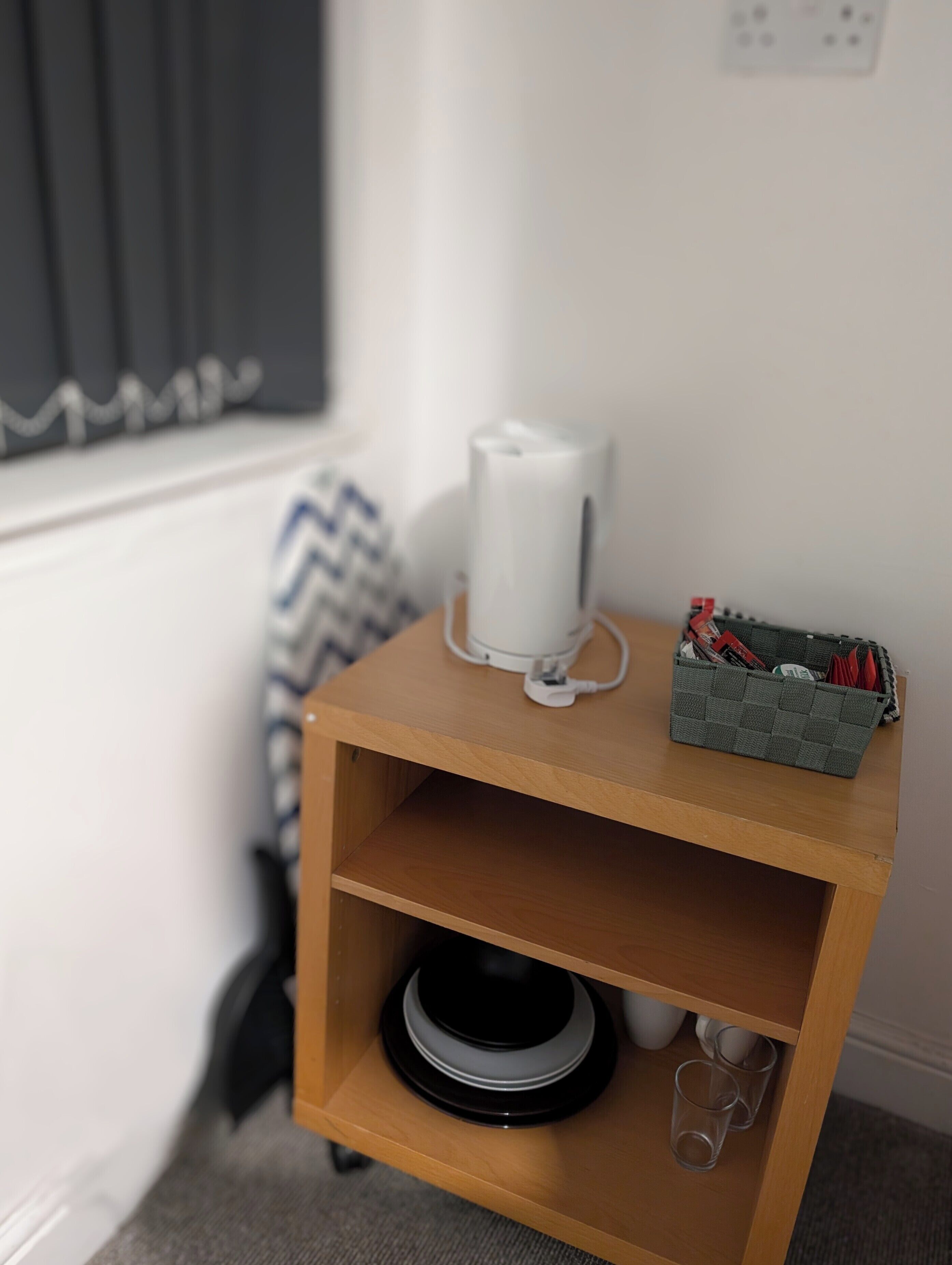 Un estante de madera pequeño con un dispositivo blanco, una cesta con objetos y un altavoz negro.