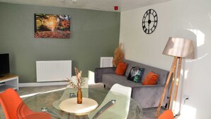 A modern living room with a glass dining table, orange chairs, a grey sofa with colorful cushions, a floor lamp, and a wall clock. - Blok 3 by SB Stayz (Blackpool)