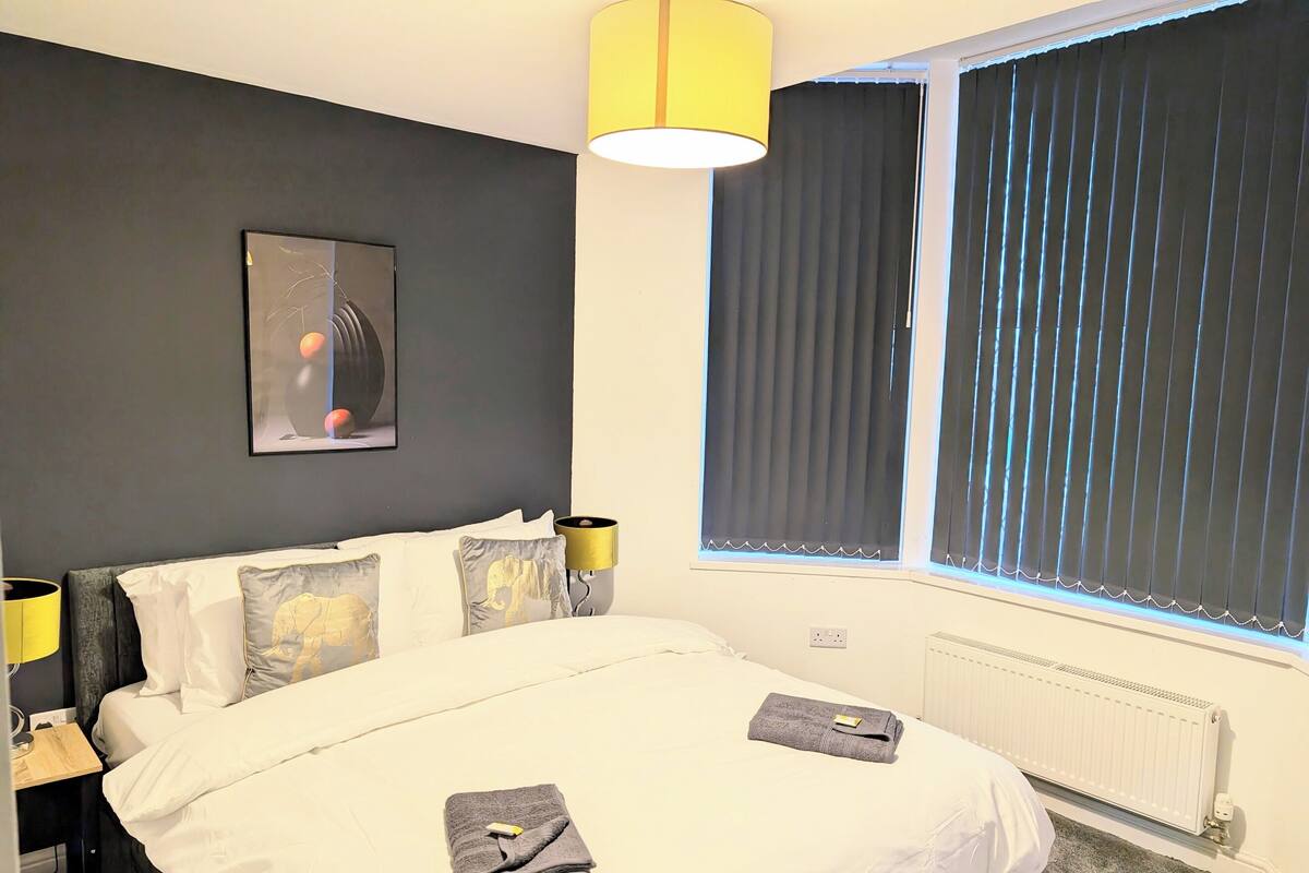 A hotel room with a bed, a bedside lamp, a window with blinds, a radiator, and a framed picture on the wall.