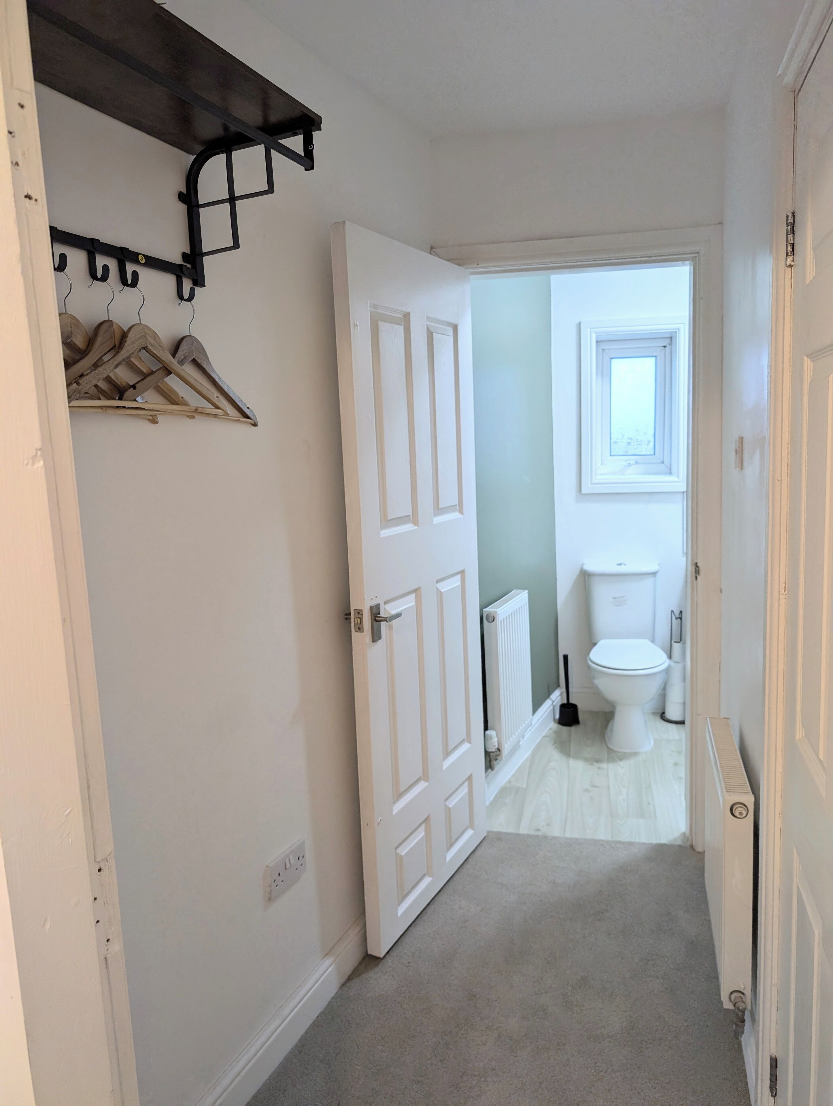 A narrow hallway with a white door leading to a bathroom. A coat rack with wooden hangers is mounted on the wall.