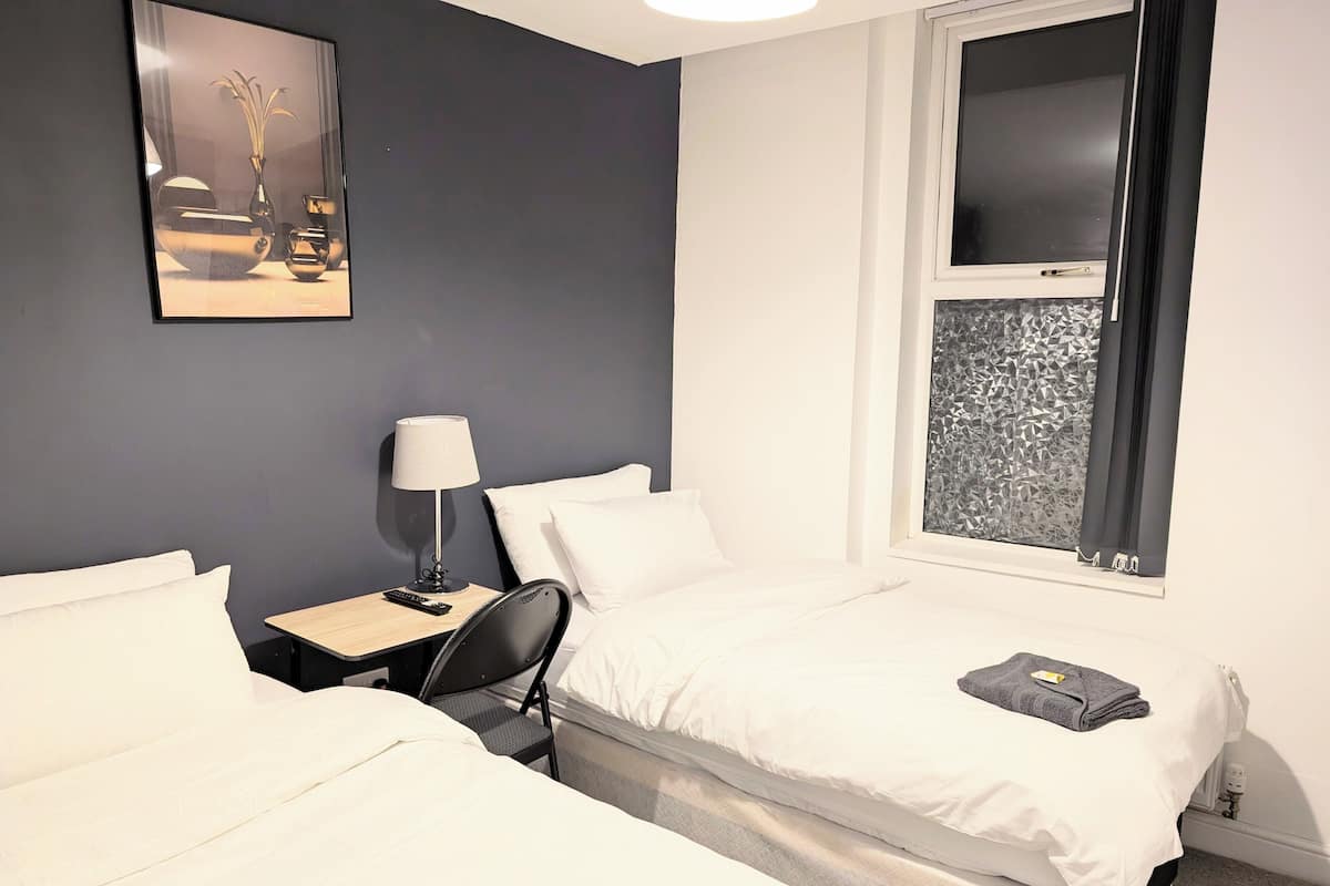 A hotel room with two beds, a desk with a lamp, a painting on the wall, and a window with a geometric pattern.