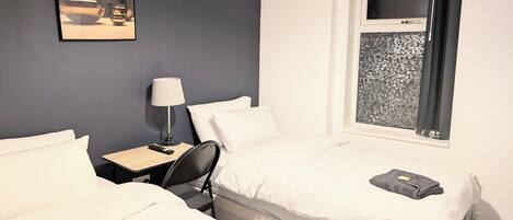 A hotel room with two beds, a desk with a lamp, a painting on the wall, and a window with a geometric pattern.