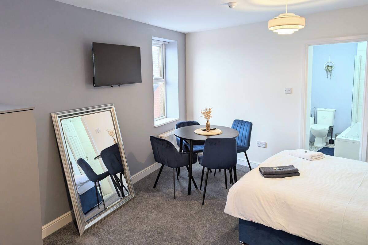 A hotel room with a bed, a round table with chairs, a television mounted on the wall, and a large mirror leaning against the wall.