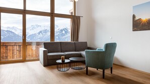 Ein modernes Wohnzimmer mit einem grauen Modulsofa, einem türkisfarbenen Sessel und zwei runden Couchtischen. Große Fenster bieten Blick auf schneebedeckte Berge.