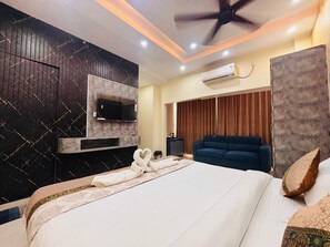 Une chambre d’hôtel avec un grand lit, un téléviseur à écran plat fixé au mur, un appareil de climatisation, un ventilateur de plafond et un canapé bleu foncé.