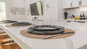 Eine moderne Küche mit weißen Arbeitsplatten, einem Essbereich mit einem langen Tisch, gedeckt für vier Personen, und einer Kücheninsel mit Spüle und Arbeitsfläche.