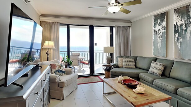 A modern living room with a sofa, a coffee table, and a TV. There is a balcony with a view of the sea.