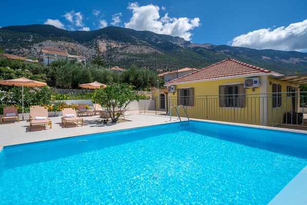 A swimming pool with lounge chairs and umbrellas, a yellow building with a red roof, and a mountainous backdrop.