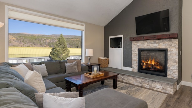 A living room with a stone fireplace, a large window with a view of a forest, and a flat-screen TV mounted above the fireplace.