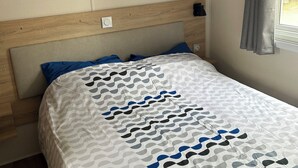 A bedroom with a wooden headboard, a bed with a patterned bedspread, a small shelf with decorative items, and a window with curtains. - Mobile home with direct beach access (Canet-en-Roussillon)