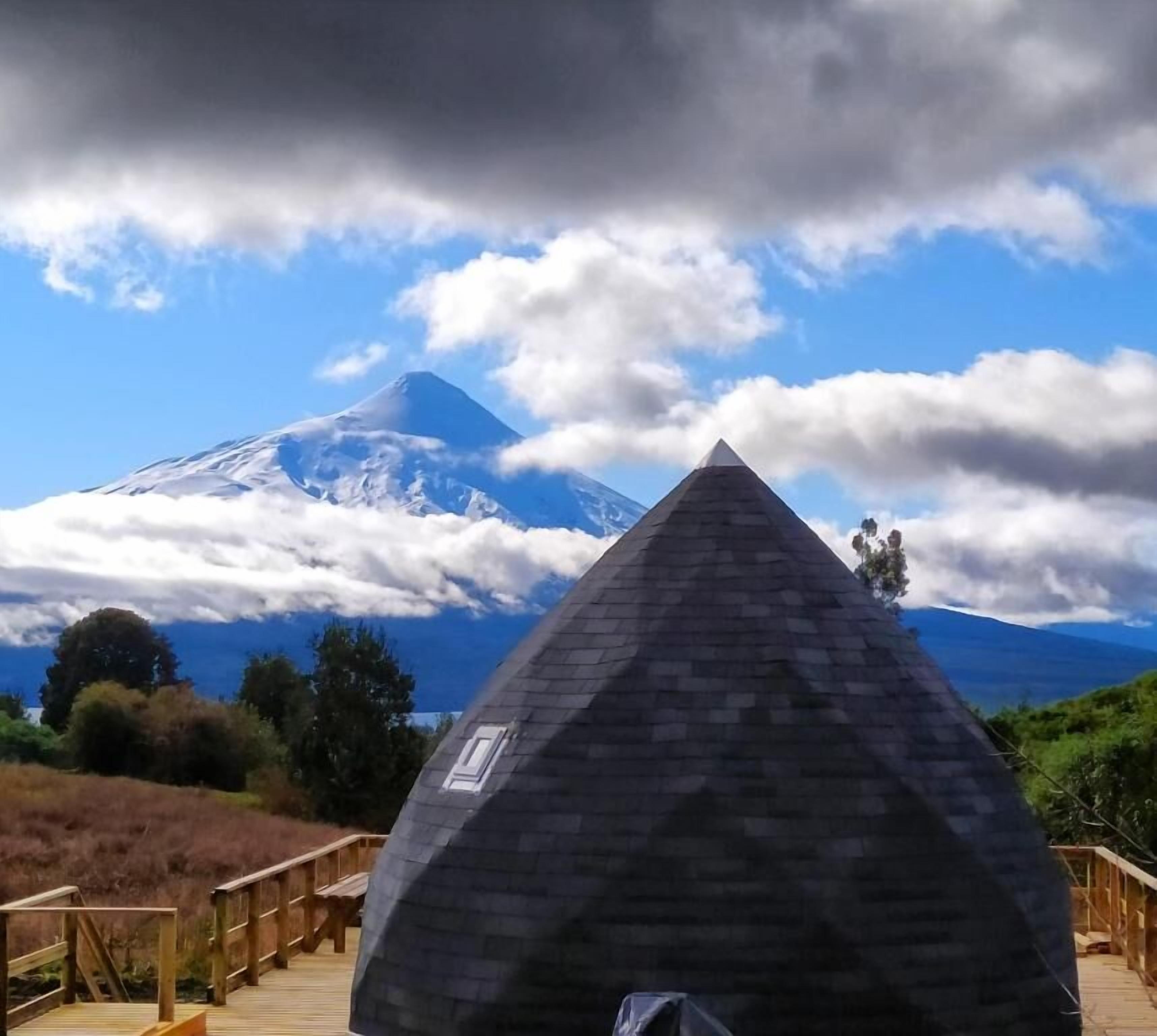 A pyramid-shaped structure with a wooden deck in the foreground and a mountain with clouds in the background.