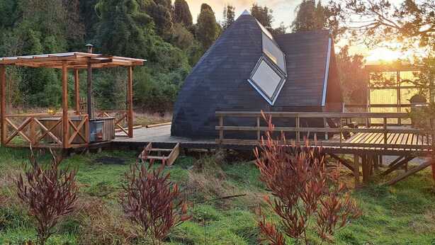 A modern house with a sloped roof and skylights, surrounded by a deck and wooden pergola, set against a forest backdrop. - Casa Pumahue  (Puerto Varas)