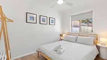 A bedroom with a bed, a wooden headboard, a bedside table with a lamp, a window with a view of a brick building, and three framed pictures on the wall.