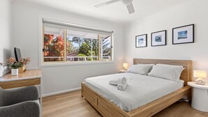 Ein modernes Schlafzimmer mit einem hölzernen Bett, einem Schreibtisch mit Computer und einem Fenster mit Herbstlaub.