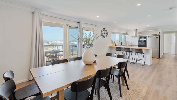 A modern kitchen with a central island, a dining area with a large table and chairs, and a view through the windows. - The Beach House 64 Neighbour Ave Goolwa Beach (Goolwa)