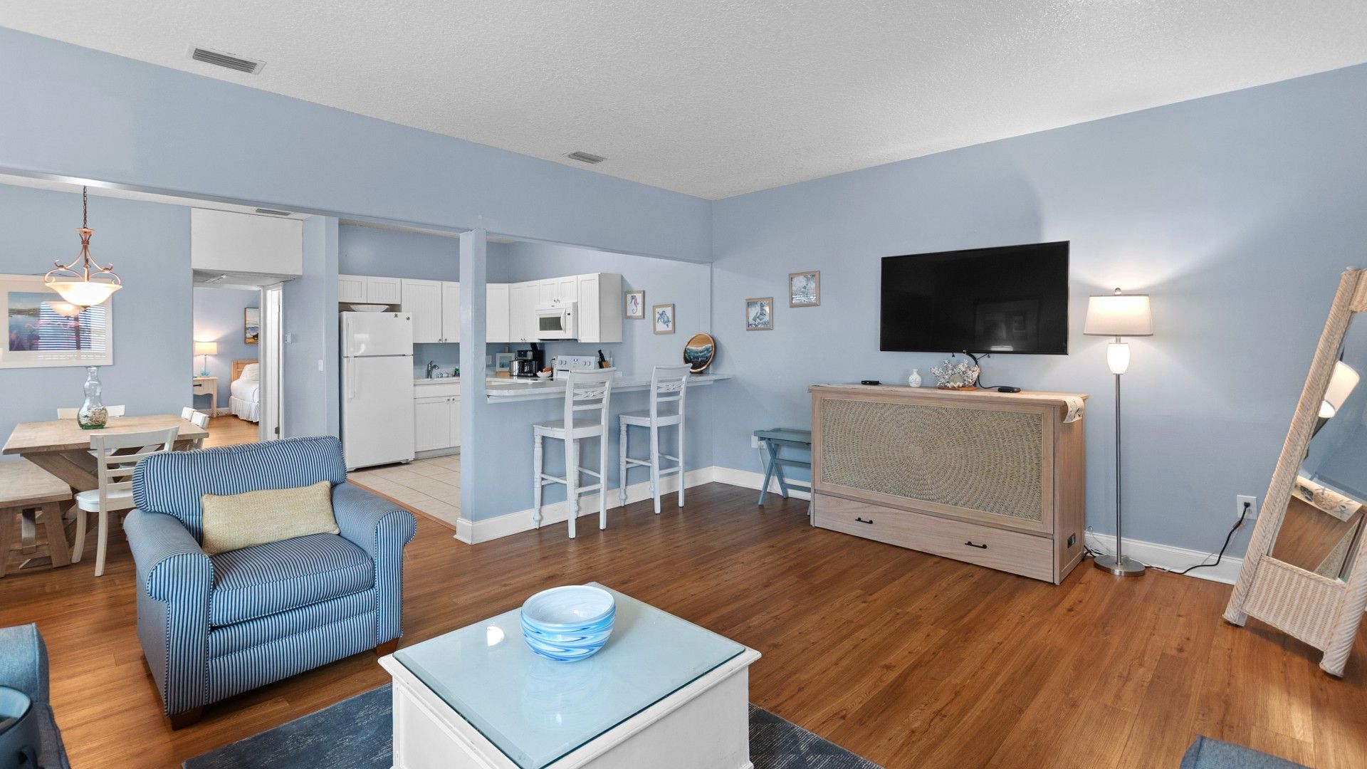 A modern living room with a sofa, coffee table, and a TV. There is a kitchen area with white cabinets and a dining table.