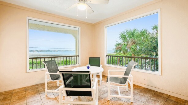 Ein Balkon mit Tisch und Stühlen, große Fenster mit Blick auf das Meer und Palmen sowie ein Deckenventilator.