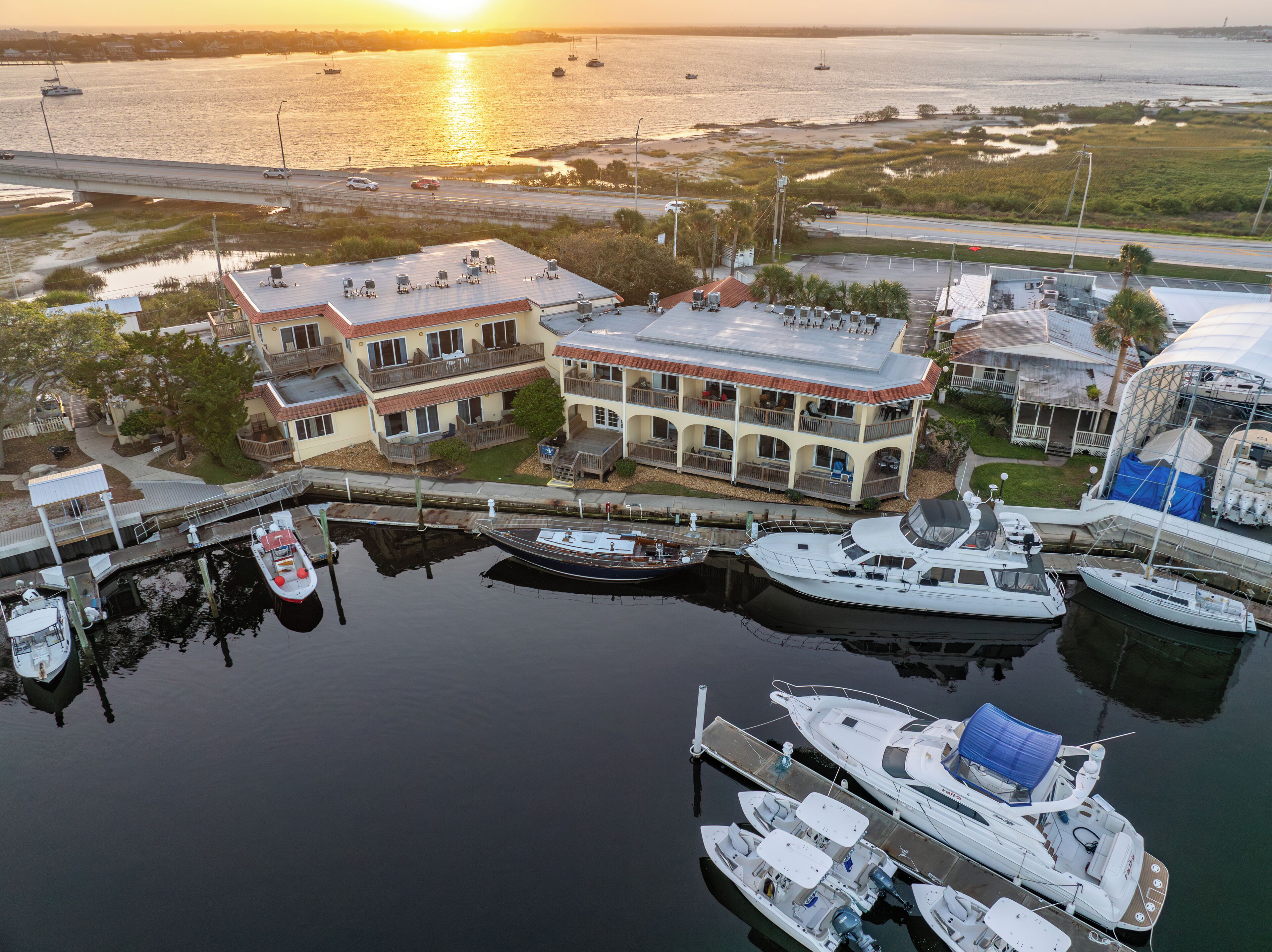 A marina with boats docked, a large building with balconies, and a sunset in the background.