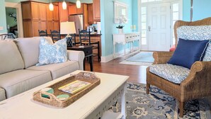 A living room with a sofa, a wicker chair, a dining table, and a kitchen area with wooden cabinets and a refrigerator. - JonBie Maisons SEA GLASS COTTAGE cart included with booking (Daufuskie Island)
