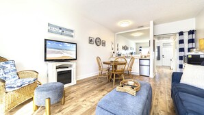 Un salon avec un téléviseur, des meubles en osier, un coin repas avec une table et des chaises, et une cuisine à l’arrière-plan.