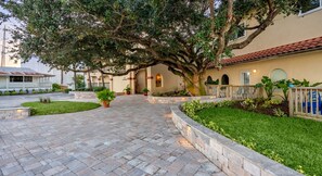 A paved pathway leading to a building with a large tree, a well-maintained lawn, and a fence with blue chairs. - Marina View Loft Studio | King Bed + Private Deck | Dogs OK (St. Augustine)