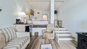 A compact living space with a kitchenette, a sofa, a chair, a small table, and a staircase leading to a bedroom.