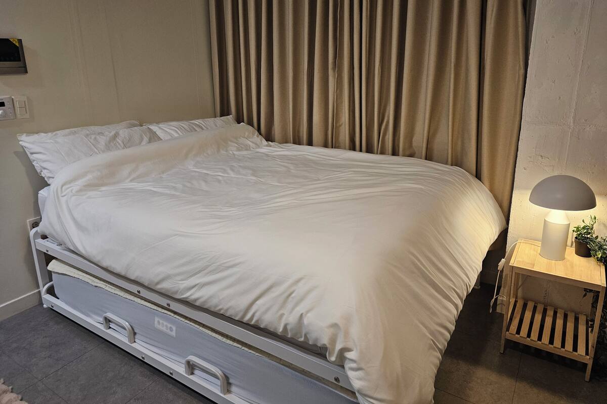 A single bed with a white mattress and pillows, a bedside table with a lamp, and a curtain covering the window.