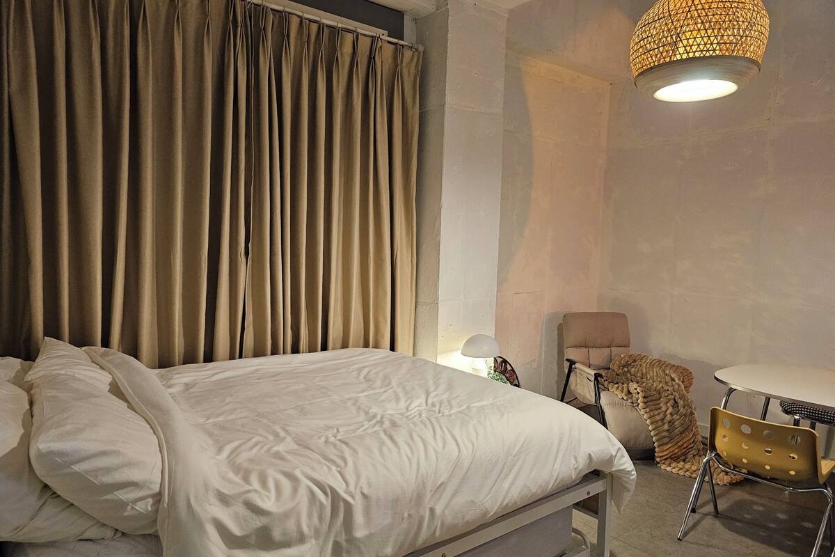 A bedroom with a bed, a chair, a small table, and a hanging light fixture.