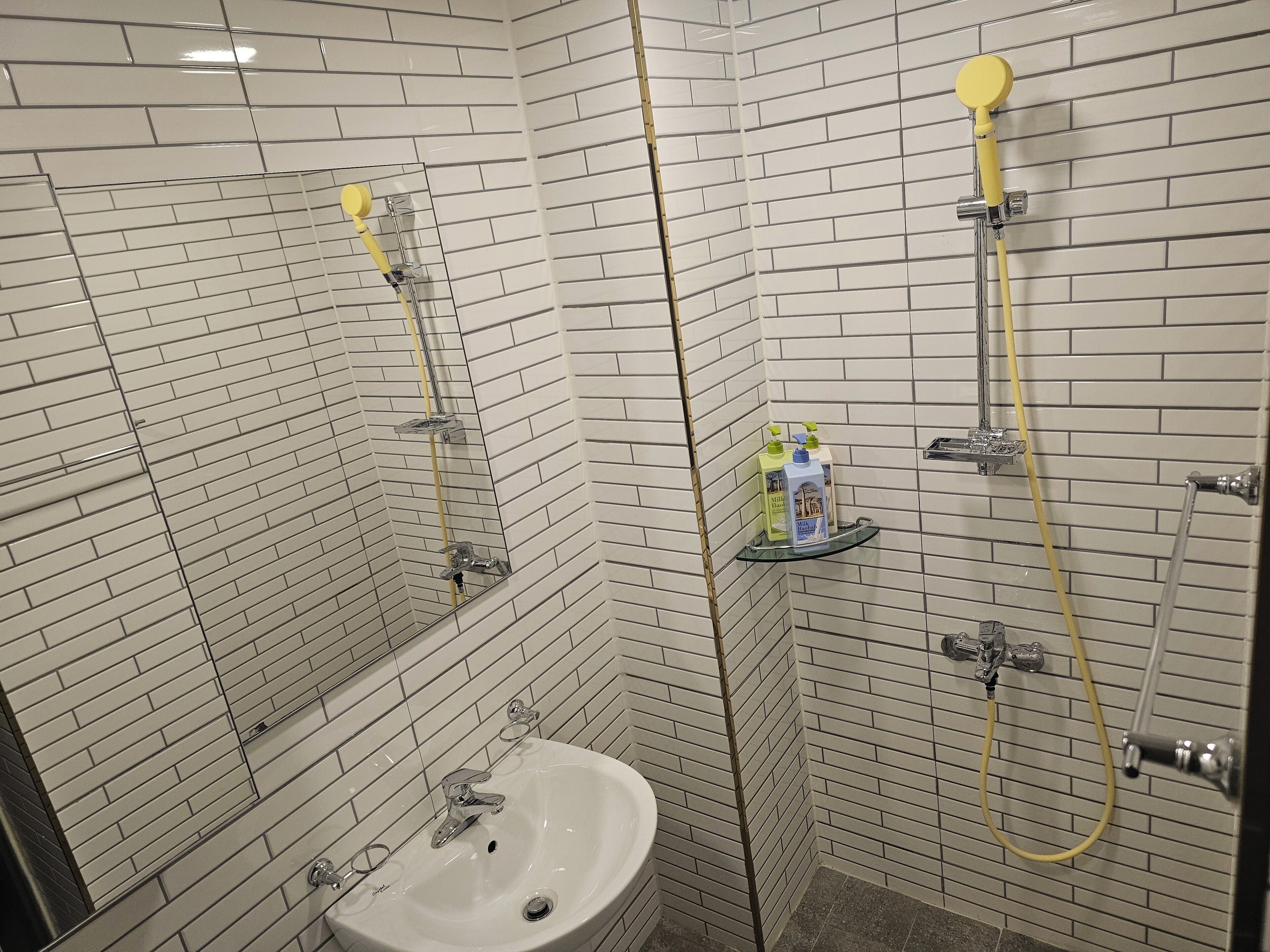 A bathroom with white tiled walls, a sink, and a shower with a yellow hose.