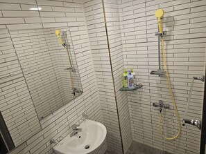 A bathroom with white tiled walls, a sink, and a shower with a yellow hose.