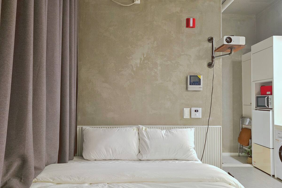 A bedroom with a bed, a wall-mounted projector, a thermostat, and a microwave.