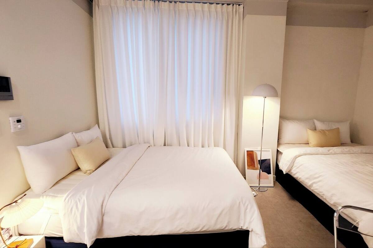 A hotel room with a large bed, a smaller bed, a bedside table, a lamp, and a window with curtains.