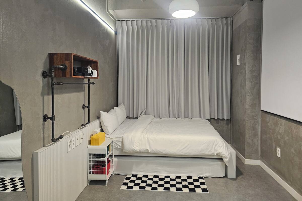 A bedroom with a bed, a radiator, a shelf with a camera, a checkered rug, and a white ceiling light.